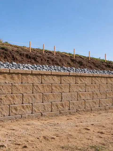 New tan segmental block retaining wall being built across a sloped San Diego backyard, with gravel behind the courses and grade stakes along the top